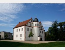 Historisches Gebäude mit weißer Fassade, roten Backsteinkanten und einem dunkelgrauen Turmdach, umgeben von einer grünen Wiese und Bäumen; daneben ein modernes, schlichtes Gebäude.
