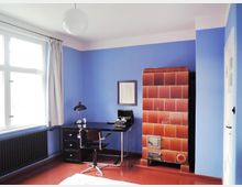 Zimmer mit blau gestrichenen Wänden, einem großen Fenster mit weißen Vorhängen und einem schwarzen Schreibtisch mit Stuhl. Rechts steht ein brauner, gefliester Kaminofen, und an der Wand hängt ein gerahmtes Bild.