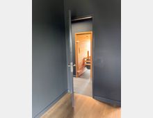 Ein Raum mit dunklen Wänden und Holzboden, in dem eine geöffnete Tür zu einem angrenzenden Bereich mit Holzverkleidung und grauem Fliesenboden führt.