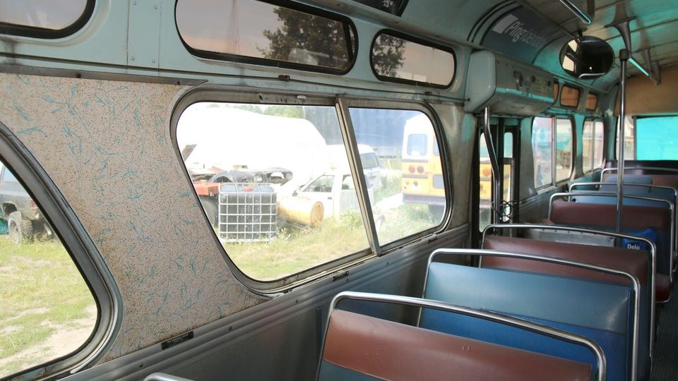 Innenraum eines alten Busses mit abgenutzten Sitzbänken in brauner und blauer Farbe. Durch die großen Fenster sind mehrere geparkte Fahrzeuge und eine grüne Wiese zu sehen.