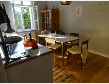 Ein helles Esszimmer mit Holzboden, einem weißen Tisch und verschiedenen Stühlen, von denen einige mit Fellauflagen versehen sind. Im Hintergrund stehen ein geöffneter Fensterrahmen, durch den Bäume sichtbar sind, und ein Holzbücherschrank, während die linke Bildseite eine Küchenzeile mit einem orangefarbenen Wasserkocher zeigt.