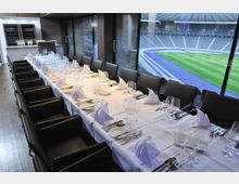 Eleganter Speiseraum mit einer langen, weiß gedeckten Tafel, die mit Gläsern, Besteck und gefalteten Servietten arrangiert ist; große Fenster bieten einen Ausblick auf ein Stadion mit einer Laufbahn und einem gepflegten Rasen.