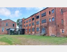 Backsteinbau mit mehreren Etagen, dunkelgrauen Fenstern und einem modernen Glas-Anbau, der zwei Gebäudeteile verbindet. Vor dem Gebäude erstreckt sich eine Grünfläche mit gepflasterten Wegen.