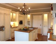 Helle, klassische Küche mit cremefarbenen Einbauschränken, einer zentralen Kücheninsel mit Holz- und Steinoberfläche, einem Kronleuchter an der Decke und Holzboden. Moderne Geräte wie ein eingebauter Kaffeeautomat und dekorative Akzente wie eine Obstschale auf der Kücheninsel sind sichtbar.