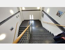 Treppenhaus in einem modernen Gebäude mit schwarzen Steinfliesen, weißen Wänden und Holzhandläufen. An den Wänden hängen mehrere kleine Bilder, und zwei schmale Fenster lassen Tageslicht herein.