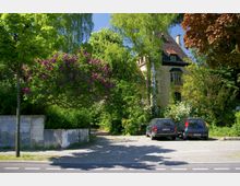 Eine historische Villa mit einem steilen Ziegeldach und dekorativen Fenstern, umgeben von dichter grüner Vegetation, darunter blühende lila und weiße Büsche. Im Vordergrund befindet sich ein kleiner Parkplatz mit zwei geparkten Autos, angrenzend an eine Straße mit Markierungen.