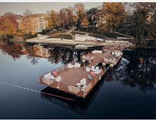Floating wooden dock platforms on a calm lake, furnished with white seating and red lounge chairs and bordered by railings. In the background, a terraced shoreline with stairways leads up to trees with autumn foliage and a large multi-story building reflected in the water.