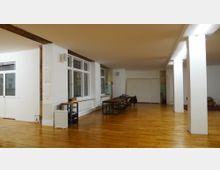 Ein großer, heller Raum mit Holzboden, weißen Wänden und deckenhohen Fenstern auf der linken Seite. In der Mitte stehen lange Holztische mit Stühlen, und der Raum wird durch mehrere weiße Säulen unterteilt.