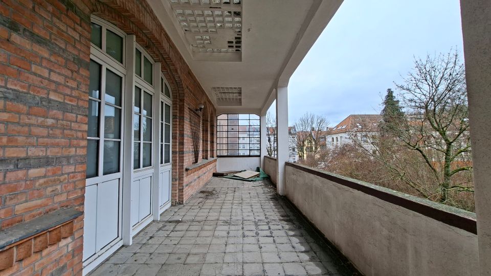 Langer, überdachter Balkon oder Laubengang an einem Backsteingebäude mit weißen Fenstern und Türen, Fliesenboden und Betondecke mit Lochmuster. Rechts verläuft eine niedrige Brüstung mit Blick auf Bäume und umliegende Wohnhäuser.