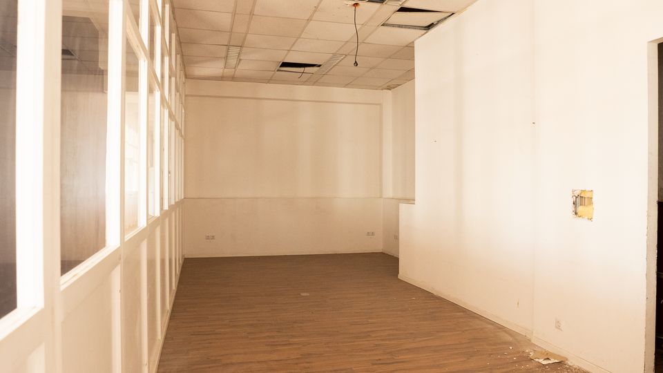 Leerer Innenraum eines ehemaligen Büro- oder Gewerberaums mit weiß gestrichenen Wänden, braunem Laminatboden und einer abgehängten Rasterdecke mit offenen Deckenfeldern und herabhängenden Kabeln. Links verläuft eine Glaswand mit Fenstern, rechts ist eine Öffnung bzw. ein Zugang in der Wand zu sehen.