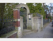 Eingangstor aus Metall mit verziertem Gitter, eingefasst von einer gemauerten Ziegel- und Betonkonstruktion mit Rundbogen und Hausnummer „7“. Im Hintergrund sind Bäume und eine gepflasterte Straße zu sehen.