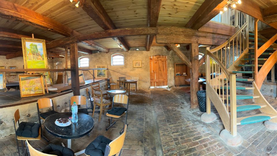 Innenraum eines rustikalen Scheunen- oder Dachbodenraums mit freiliegenden Holzbalken, Backsteinboden und steinernen Wänden, teils als Atelier mit Staffelei und gerahmten Gemälden genutzt. Rechts führt eine Holztreppe mit Geländer zu einer Galerieebene; im Vordergrund stehen ein runder Tisch mit Stühlen sowie eine Couch.