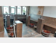 Raum mit mehreren kachelverkleideten Sitzbänken und Säulen, die mit Wasserhähnen ausgestattet sind, sowie Holzeimern auf dem Boden. Im Hintergrund befinden sich drei bogenförmige Fenster mit Pflanzendekoration und Laternen.