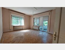 Leerer Raum mit Parkettboden, großen Fenstern und einer Balkontür, die mit Jalousien abgedeckt sind. Die Wände sind hell beige, und unter den Fenstern befinden sich Heizkörper.