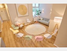 Heller Raum mit hellem Holzfußboden, in dem eine Sitzgruppe aus bunten Stühlen kreisförmig um einen dekorativen runden Teppich angeordnet ist. Im Hintergrund stehen ein graues Sofa, Pflanzen und dekorative Accessoires vor großen Fenstern mit blickdichten Rollläden.