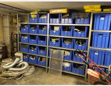 Werkstattraum mit mehreren Metallregalen, die mit blauen Kunststoffkisten gefüllt sind. Im Vordergrund liegen Kabelrollen und ein flexibler Lüftungsschlauch, daneben stehen Werkzeuge und weitere Geräte. Die Wände sind aus unverputztem Mauerwerk.