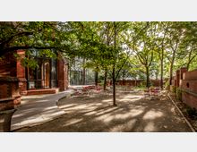 Außenhof mit rotbraunem Backsteingebäude und großen Glasfenstern auf der linken Seite, umgeben von Bäumen. Der Hof ist mit runden Tischen und roten Stühlen möbliert, daneben stehen eine gepflasterte Gehwegführung und eine Mauer aus Backstein als Begrenzung.