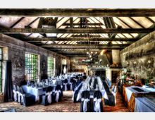 Ein rustikaler Veranstaltungsraum mit Holzbalkendecke, großen Fenstern mit grünem Ausblick und dekorierten Esstischen mit weißen Tischdecken und schwarzen Stuhlbezügen. Im Hintergrund befindet sich ein Kamin, und die Wände sind mit Reliefs und Dekorationen geschmückt.
