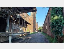 Ansicht eines ehemaligen Fabrikgeländes mit Backsteinfassaden. Links ein Gerüst an einem Gebäude mit der Aufschrift „LSR →“, rechts eine mit Efeu bewachsene Fassade, und in der Mitte ein mit Kopfsteinpflaster ausgelegter Weg, der zwischen den Gebäuden in die Tiefe führt.