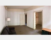 Wohnzimmer in einem älteren Gebäude mit weißen Wänden, braunem Teppichboden und einfacher Einrichtung. Es sind eine schwarze Couch, eine Stehlampe, eine weiße Zimmertür und zwei offene Durchgänge sichtbar, die zu anderen Räumen führen.