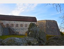 Historisches Gebäudekomplex mit Steinmauer und roten Dachziegeln, im Hintergrund ein massiver Rundturm aus Naturstein; im Vordergrund größere Felsen und leicht bewachsene Rasenfläche. Der Himmel ist klar mit einigen Wolken.