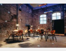 Industrie-inspiriertes Interieur mit rauen Ziegelwänden, hohen Decken und großen Fenstern. Mehrere Holztische und Stühle sind im Raum verteilt, beleuchtet von hängenden Lampen; im Hintergrund hängt ein schwarzer Vorhang.
