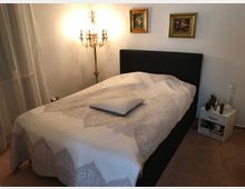 Schlafzimmer mit einem zentralen Doppelbett mit gemustertem Überwurf und Kissen. Im Hintergrund ein dekorativer Kronleuchter an der Wand sowie zwei gerahmte Blumenbilder, daneben ein Nachttisch mit Lampe und persönlichen Gegenständen.