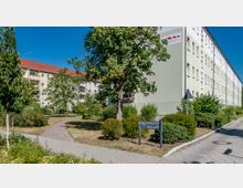 Mehrstöckige Wohngebäude mit hellem Putz und roten Dächern, umgeben von einem begrünten Innenhof mit Bäumen, Büschen und einem gepflasterten Gehweg. Im Vordergrund ein Schild mit der Aufschrift „Rosa-Luxemburg-Straße 38-46“.