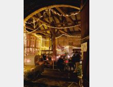 Ein rustikaler Innenraum mit Holz- und Ziegelstrukturen, dekoriert mit warmen Lichterketten. Im Vordergrund sitzen Personen an einem Tisch, während im Hintergrund dekorative Gegenstände wie ein Kinderfahrrad und ein gelber Kleintransporter sichtbar sind.