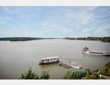 Flusslandschaft mit einer Anlegestelle, an der ein kleines Passagierschiff anlegt; im Vordergrund Büsche und Schilf, im Hintergrund bewaldete Ufer und eine Ansiedlung.
