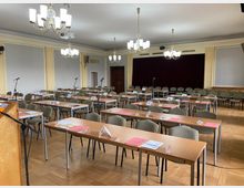 Ein Konferenzraum mit mehreren Reihen von Holztischen und gepolsterten Stühlen, auf den Tischen liegen Namensschilder und Dokumente. Der Raum hat eine Bühne mit rotem Vorhang im Hintergrund, helle Wände, Parkettboden und mehrere Kronleuchter an der Decke.