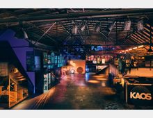 Eine weitläufige, industriell gestaltete Halle mit hohen Decken, freiliegenden Metallträgern und bunter Beleuchtung. Im Innenraum befinden sich mehrere Ebenen mit Glasfenstern, Treppen und diversen dekorativen Elementen, darunter ein Schild mit der Aufschrift „KAOS“.