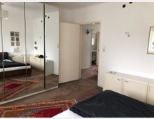 Schlafzimmer mit modernem Design, ausgestattet mit einem großen Spiegel-Kleiderschrank, einem Doppelbett, Teppichen in Rot- und Goldtönen sowie weißen Möbeln. Im Hintergrund ist eine offene Tür mit Blick auf ein angrenzendes Badezimmer zu sehen.