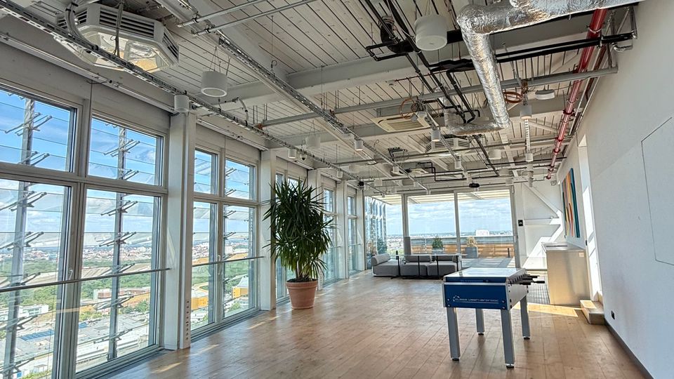 Open-plan modern office lounge with light wood floors, floor-to-ceiling windows along one wall, and an exposed industrial ceiling with visible ducts, pipes, and wiring. The space includes a potted plant, seating area, and a foosball table, with city views through the windows.