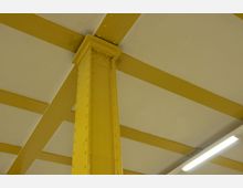 Gelbe Stahlträger und eine gelbe Stütze mit Nieten, die eine weiße, segmentierte Decke tragen; rechts ist eine helle Leuchtstoffröhre sichtbar.