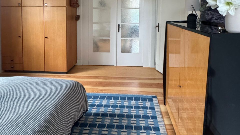 Schlafzimmer mit Holzboden und blau gemustertem Teppich, links ein Bett sowie ein großer Holzkleiderschrank unter einer Hängelampe. Hinten stehen weiße Doppeltüren mit Milchglas, rechts eine Kommode und eine offene Tür zu einem Nebenraum.