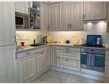 Küche mit hellen Holzmöbeln, grauer Arbeitsplatte und gefliester Rückwand. Zu sehen sind ein eingebauter Backofen mit Gasherd, eine Mikrowelle, ein Spülbecken mit Geschirrständer sowie verschiedene Küchengeräte und Dekorationsgegenstände.