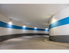 Gewundene Zufahrtsrampe einer Tiefgarage mit weißen Wänden, einem durchgehenden blauen Streifen und grauen Sockeln. Die Fläche ist aus Beton, und die Beleuchtung erfolgt durch an der Wand montierte Leuchten.