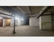 Ein leerer Kellerraum mit weißen Backsteinwänden, sichtbaren Rohren und Kabeln an der Decke. Der Boden ist betoniert, und die Beleuchtung erfolgt durch Neonröhren entlang der Decke.