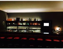 Eine Bar mit einer dunklen Theke und roten Barhockern, dahinter ein Regal mit Flaschen, Gläsern und Getränken. Der Raum ist dezent beleuchtet, mit einem kleinen Lichtakzent auf der rechten Seite.