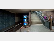 U-Bahn-Station mit Gleis auf der linken Seite, einer blau gefliesten Wand und drei Monitoren. Rechts führen eine Treppe und eine Rolltreppe zu einem höheren Niveau, während Personen diese hinaufgehen.