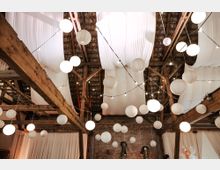 Rustikale Scheune mit Holzbalken und einer dekorativen Decke aus weißen Stoffbahnen. Zahlreiche runde Papierlaternen und Lichterketten hängen von der Decke und schaffen eine festliche Atmosphäre.