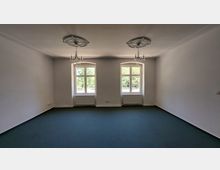 Ein leerer Raum mit weiß gestrichenen Wänden, zwei Fenstern mit weißen Holzrahmen, die nach außen auf eine grüne Umgebung blicken, und einem dunkelgrünen Teppichboden. An der Decke befinden sich zwei dekorative Stuckrosetten mit einfachen Kronleuchtern, und unter den Fenstern sind weiße Heizkörper angebracht.