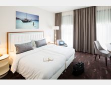 Modernes Hotelzimmer mit Doppelbett, weißen Bezügen und grauen Kissen. Ein großes Fenster mit halbtransparenter Gardine und blickdichten Vorhängen, ein Sessel mit Laptop, ein kleiner runder Tisch und eine Reisetasche auf dem Teppich ergänzen den Raum. Wanddekoration in Form eines Bildes mit Strandmotiv.