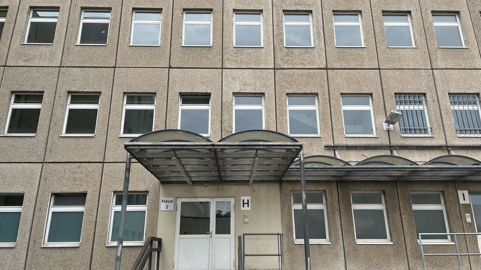 Mehrstöckiges Verwaltungs- oder Bürogebäude mit grauer Betonfassade und regelmäßig angeordneten rechteckigen Fenstern. Im Vordergrund befindet sich ein Eingang mit Treppe, weißer Doppeltür und einem gebogenen Metall-Vordach.