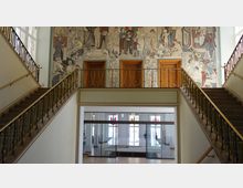 Treppenhaus eines öffentlichen Gebäudes mit symmetrisch angeordneten Treppen, Geländern mit goldfarbenen Verzierungen und einem Wandmosaik, das figürliche Szenen zeigt. Im Hintergrund befinden sich drei Holztüren auf der oberen Etage und große Fenster mit bunten Details im Erdgeschoss.