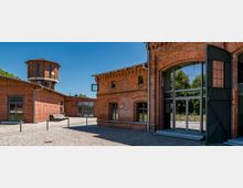 Innenhof eines umgebauten Industriekomplexes aus rotem Backstein mit Kopfsteinpflaster, großen Glasfenstern und einem modernen Eingang mit dunklen Metallrahmen. Im Hintergrund steht ein achteckiger Backsteinturm mit Uhr, flankiert von weiteren niedrigen Backsteingebäuden.