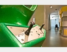 Nahaufnahme eines grünen Steuerpults mit zwei Joysticks und Beschriftungen wie "Rotate Left" und "Thrust". Im Hintergrund befindet sich ein heller Innenraum, in dem zwei Personen auf Sitzwürfeln vor einem Fenster sitzen.