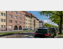 Straßenansicht mit einer Reihe von mehrstöckigen Wohngebäuden im Stil des frühen 20. Jahrhunderts, mit gelben und roten Fassaden sowie mehreren rechteckigen Fenstern. Im Vordergrund parken Autos entlang eines Gehwegs, der von Bäumen gesäumt ist.