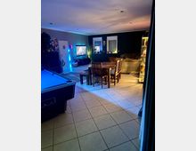 Großer Wohn- und Essbereich mit Fliesenboden, Esstisch mit Stühlen in der Mitte, Sofa-Ecke und beleuchteter Vitrine rechts. Links steht ein Billardtisch, an der Wand hängen ein Fernseher und Dekoration; farbige LED-Beleuchtung und zwei Fenster prägen den Raum.